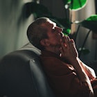 A man is sitting on a dark couch alone. He looks unhappy, he holds his hands to his face and closes ...