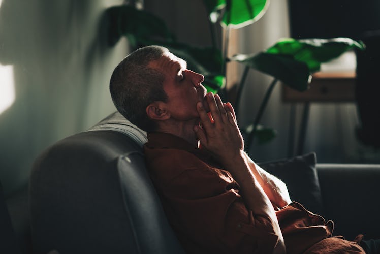 A man is sitting on a dark couch alone. He looks unhappy, he holds his hands to his face and closes ...