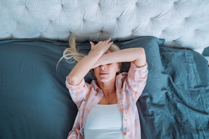 Struggling blond woman with a wild migraine, lying on bed in light bedroom. Top view