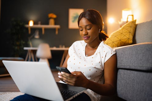 A young woman of Black ethnicity using mobile phone while working on laptop from home