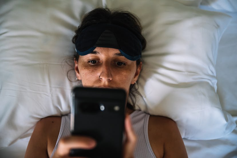 Young woman lying on a bed with a mask on her forehead and looking at her cell phone.