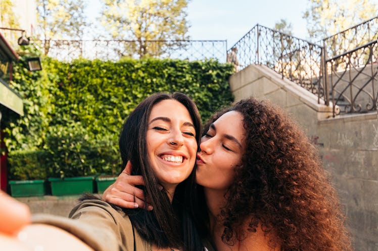 two young women pose for a selfie together as one kisses the other on the cheek, as they consider wh...