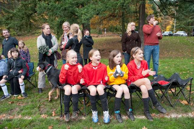 young soccer players sitting on chairs on sideline, parents and families talking and cheering in the...