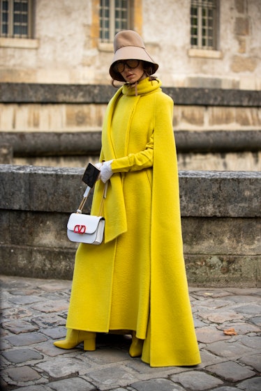 Nadia Lee Cohen, wearing a yellow long coat, white Valentino bag, beige hat and white gloves, is see...