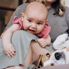 A mom sitting on a couch with her baby in her lap rubs their pet dog's belly.