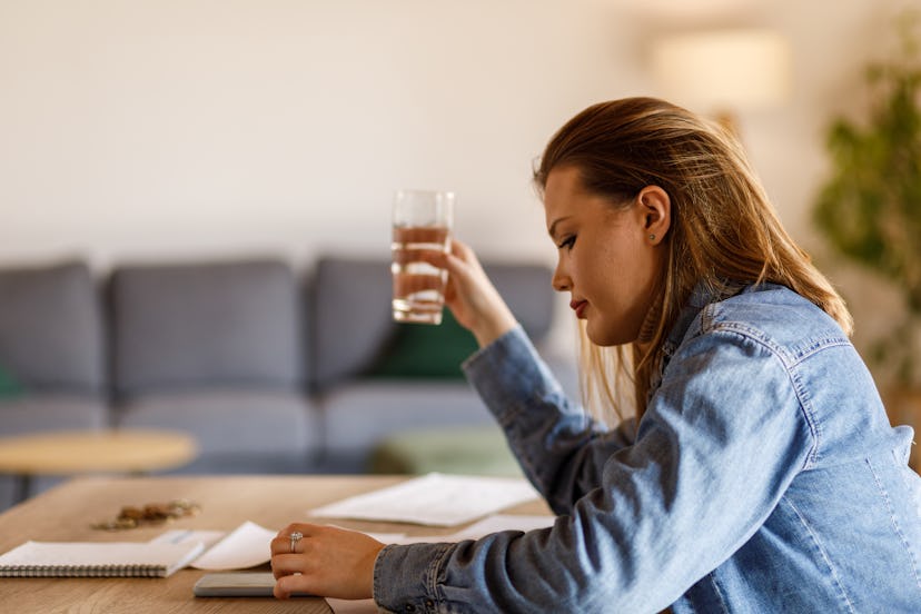 Profile view of diligent young woman sitting at dining table, drinking a glass of water and sorting ...