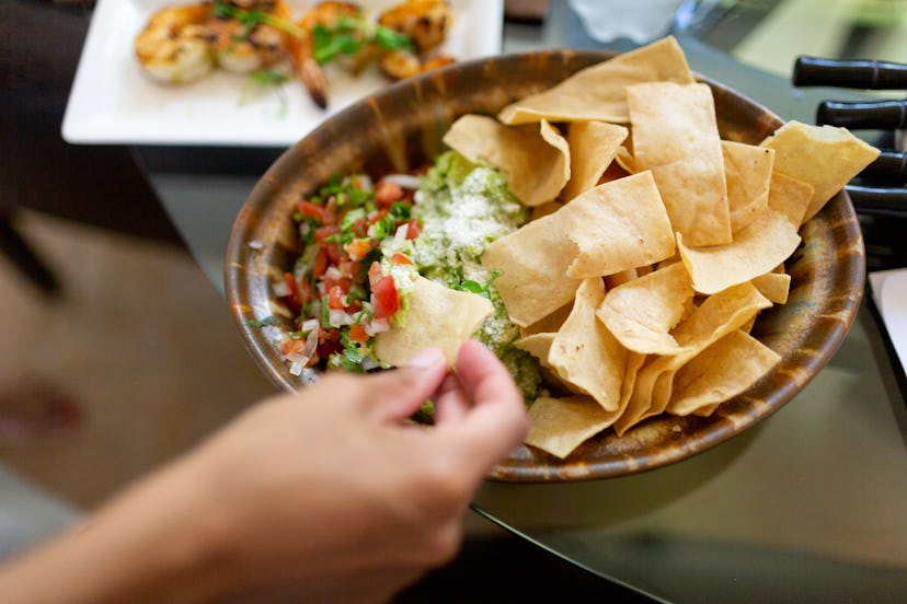 A hand reaches into a brown bowl of tortilla chips with guacamole and pico de gallo.