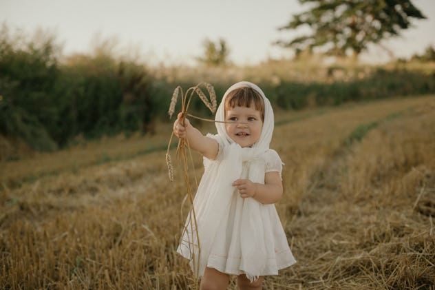 An outdoor summer portrait of an adorable toddler girl with brown hair and bangs in a cute white dre...