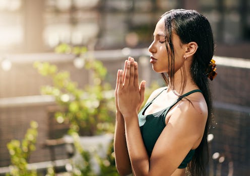 A beautiful young woman with long black hair sits doing yoga, holding her hands together with her ey...