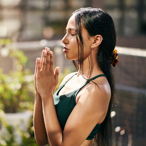 A beautiful young woman with long black hair sits doing yoga, holding her hands together with her ey...