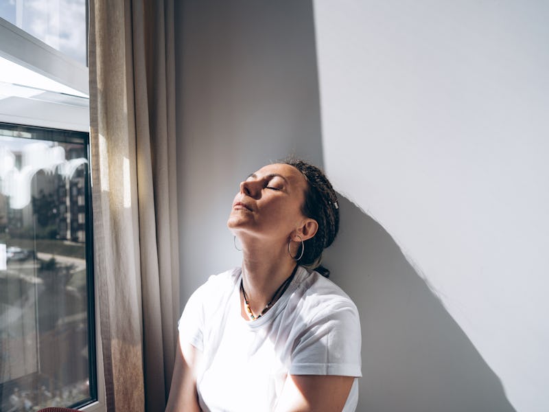 Portrait of a dreadlock braid woman sitting by the open window at the white wall. Concept of depress...