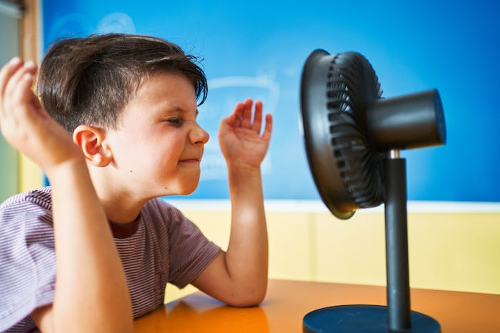 Boy refreshing his face and having fun while sitting in front of a small electric fan in the classro...