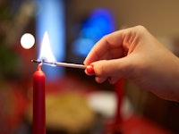 A hand carefully lights a red candle, a symbol of warmth and festivity, against a soft-focus backgro...