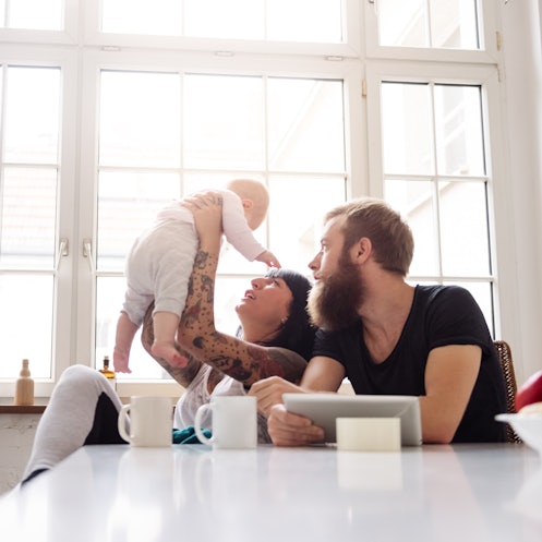 Young tattoed mother and father with newborn baby sitting in their kitchen and having fun together