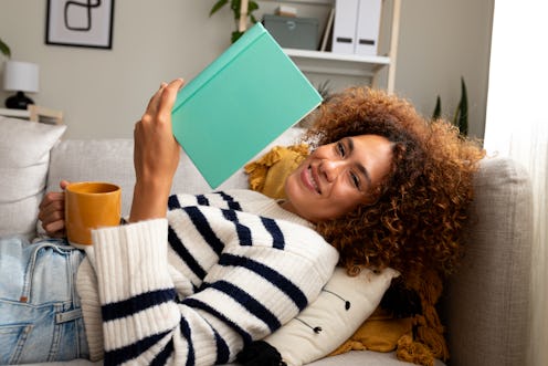 Young multiracial woman reading a book at home, drinking tea lying down on the sofa in cozy living r...