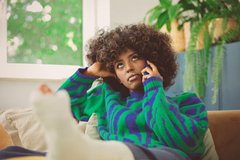 Worried afro american young woman wearing sweater sitting on sofa at home with broken leg in a plast...