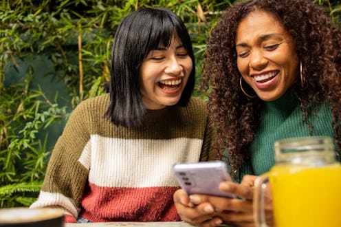 Two young women sitting at the coffee shop and using mobile phone. They are positive, smiling emotio...