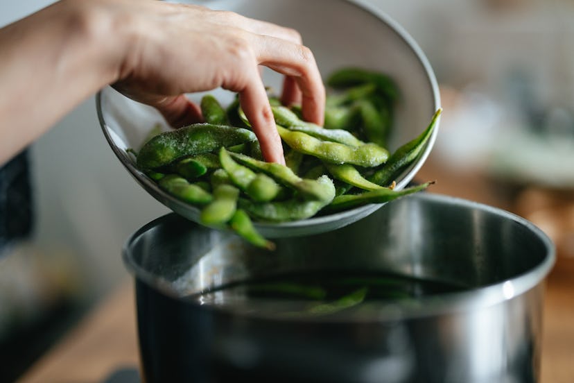 An anonymous Asian female chef standing at the gas stove and putting edamame beans into boiling wate...