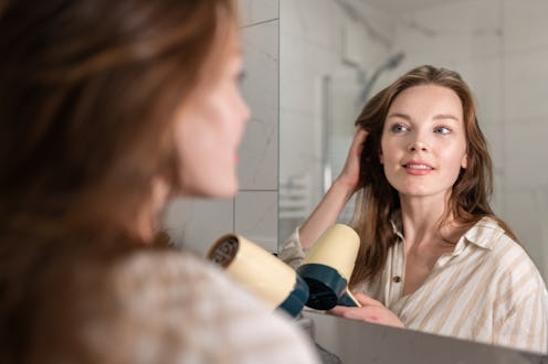 A woman is fixing her hair in the bathroom, drying it with an electric hair dryer and straightening ...
