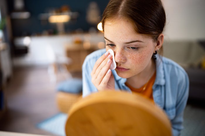 Teenage girl cleaning her face with cotton pad in front of the mirror.