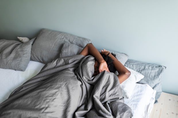 Depressed young adult black woman with headache lying in bed and hiding under blanket