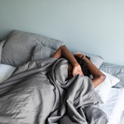 Depressed young adult black woman with headache lying in bed and hiding under blanket
