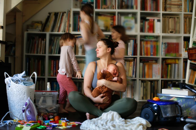 Tired, burnt-out mother with baby in her arms and out of focus children run around. Mother is sittin...
