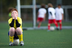 Young boy sitting on a soccer ball on a soccer pitch with his hands covering his face as three kids ...