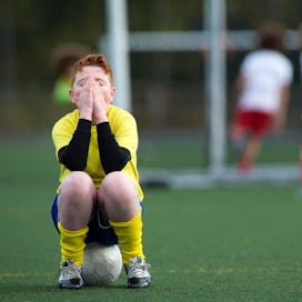 Young boy sitting on a soccer ball on a soccer pitch with his hands covering his face as three kids ...