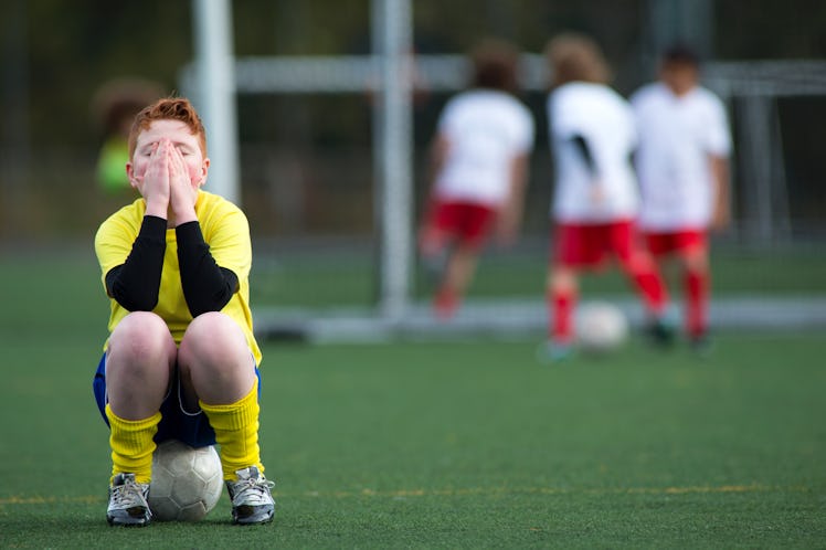 Young boy sitting on a soccer ball on a soccer pitch with his hands covering his face as three kids ...