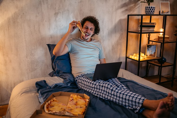 Handsome young man eating pizza and watching a movie on the laptop in the bed at night in his bedroo...