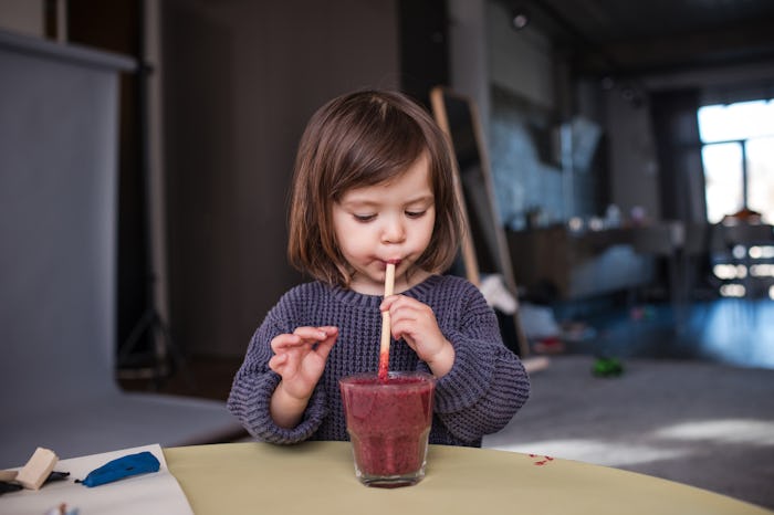 View of a beautiful baby drinking fruit smoothie from a glass with a straw. Room on the background.