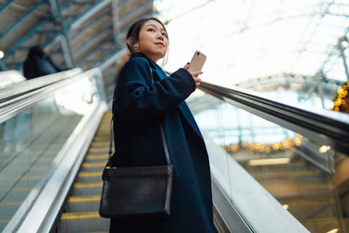 Young Asian woman with smartphone, riding on escalator, commuting to work in the city. Business trav...