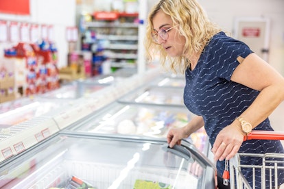 A woman in a grocery store is standing by a freezer and looking for frozen chicken.