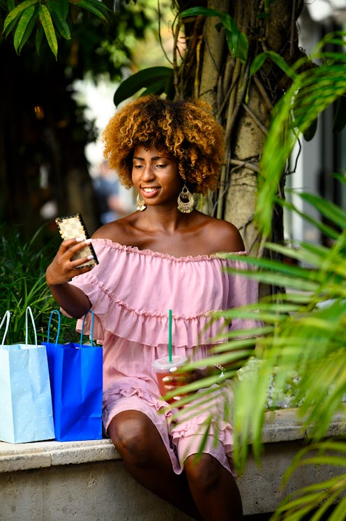 Smiling young woman holding drink while sitting against tree. Portrait of a female is with shopping ...