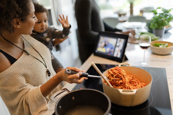 Mother holding baby daughter and cooking spaghetti dinner at stove while video chatting with family ...