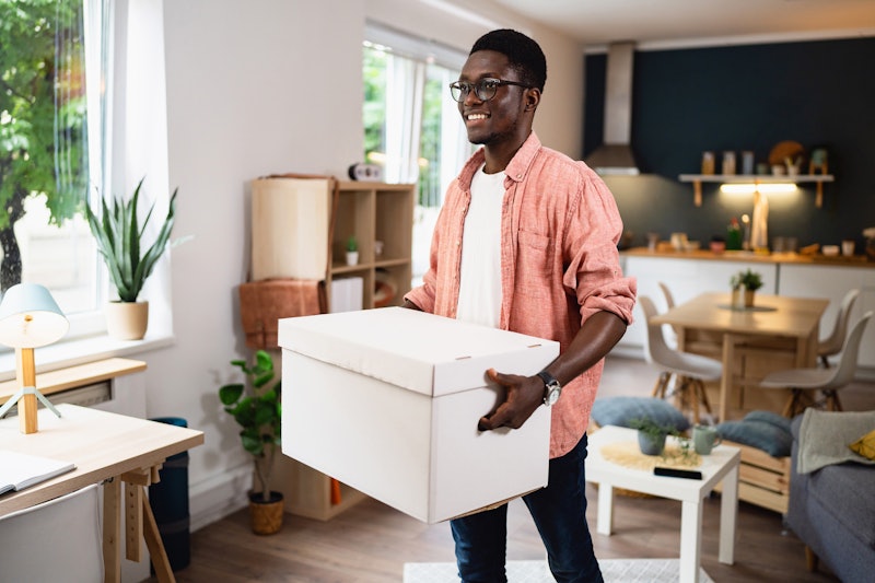 Young African-American man, carrying a cardboard box, while moving in the apartment or college dorm