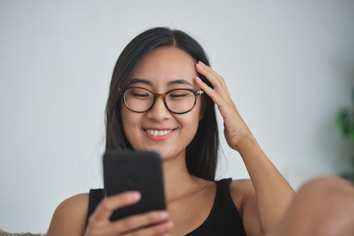 Online dating. Happy relaxed Asian girl looking at smartphone screen online messaging, chatting with...