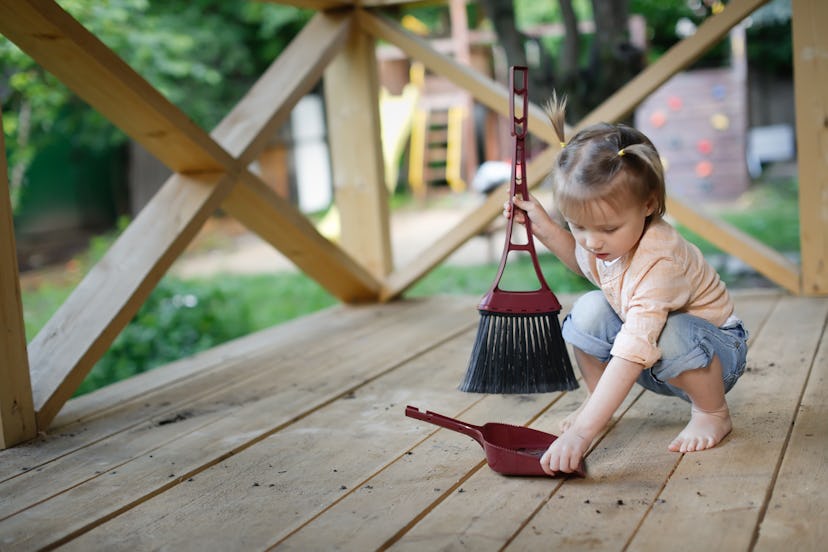 Child toddler with large broom sweeps wooden floor on veranda in summer. Developmental benefits of p...