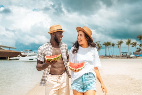 A cheerful Asian woman and a Black man savor slices of watermelon against a backdrop of palm trees a...