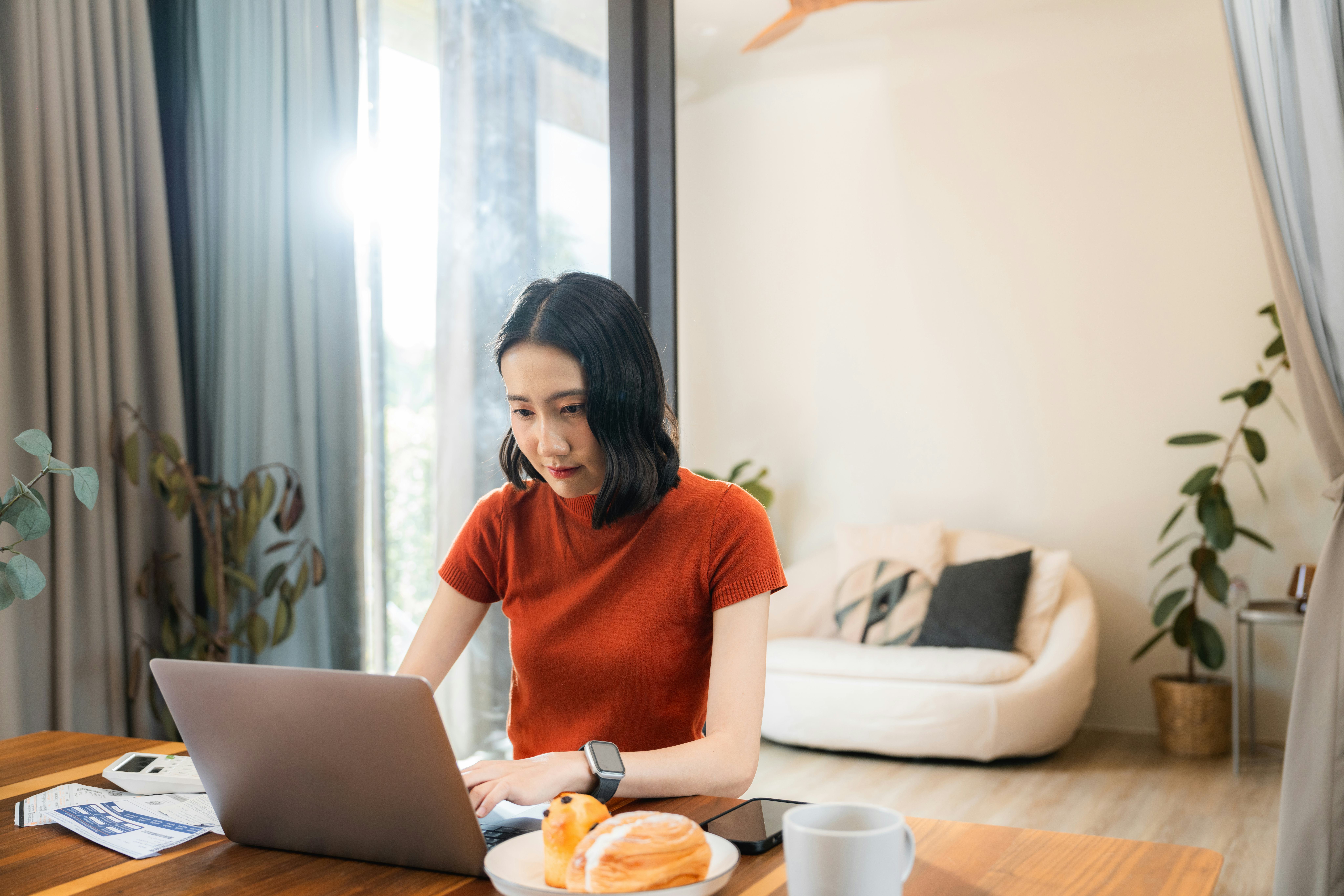 Young Asian woman working from home in the morning, typing on laptop, sitting at the desk with coffe...