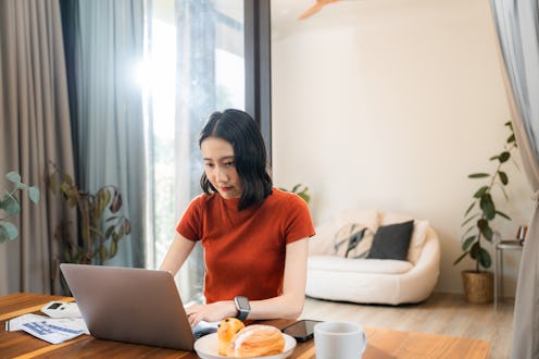 Young Asian woman working from home in the morning, typing on laptop, sitting at the desk with coffe...