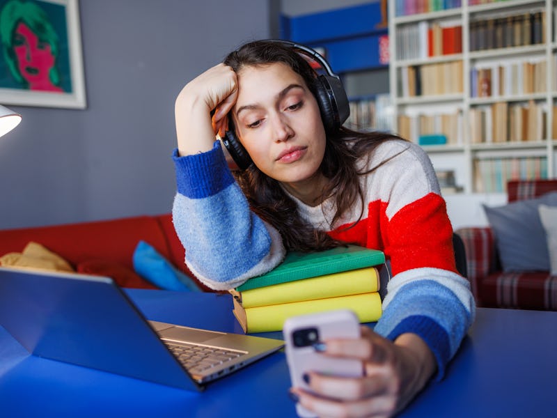 Young woman wearing headphones and looking at her phone, feeling distracted while studying at home w...