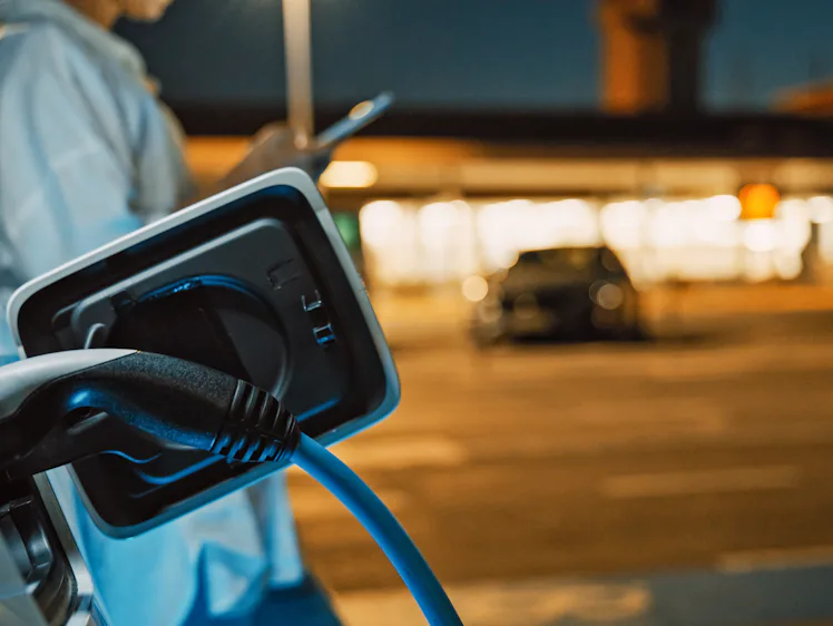 A mid-adult Caucasian woman is engaged with her smartphone while her car charges at a public electri...