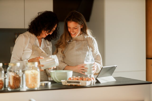 A mother and daughter stand together in a kitchen, preparing food while consulting a recipe on a dig...