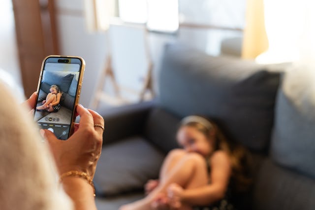 Horizontal close-up photo of a mom using phone to take a picture of her little girl on the sofa.