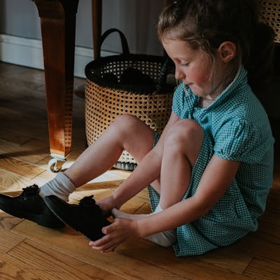 A little girl sits on the floor. She is wearing a school uniform dress. She pulls on a pair of black patent school shoes. A routine board can help kids become more independent faster.