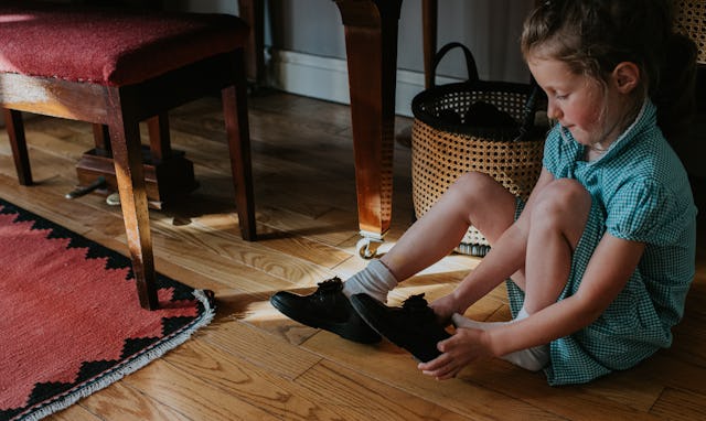 A little girl sits on the floor. She is wearing a school uniform dress. She pulls on a pair of black...