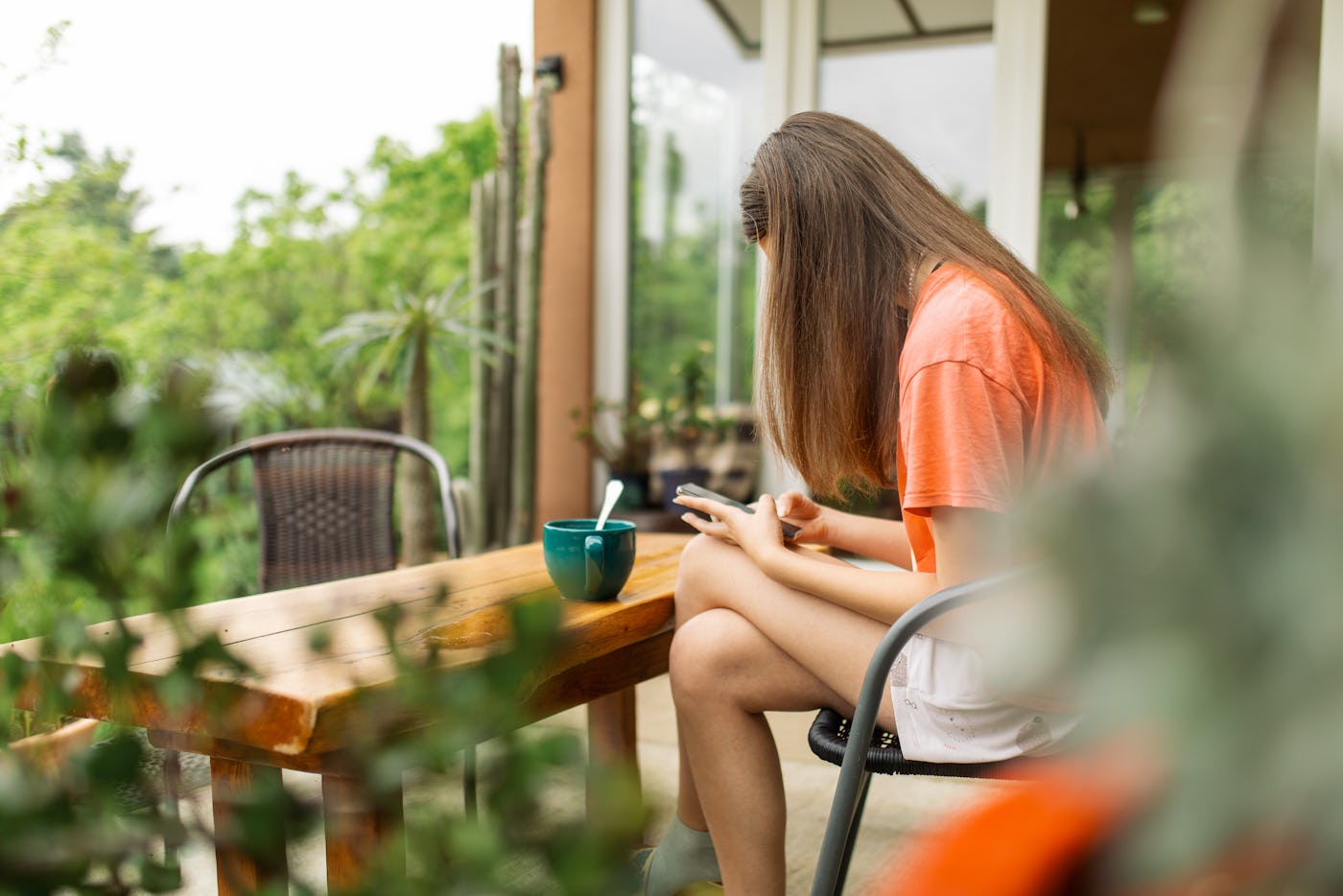 Teenage girl sitting on patio texting on her phone, looking peaceful. It's springtime, she is surrounded with plants.