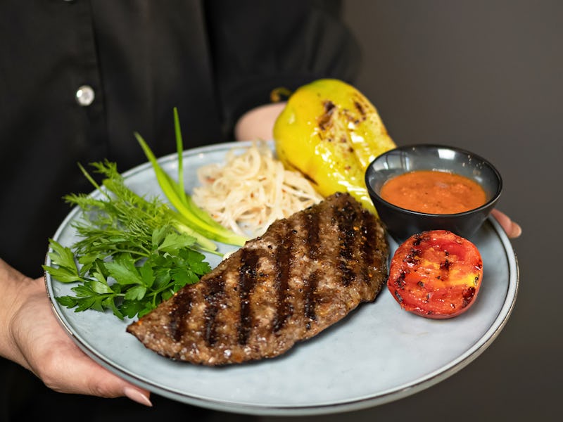 Waiter in black uniform serves lunch. Grilled meat steak with vegetables, herbs and sauce on plate i...